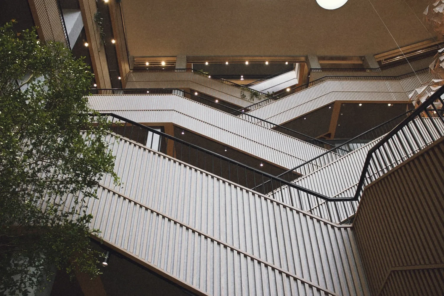 Interior view of a multilevel hall in Arbejdernes Landsbank with white railings, wooden accents, and architectural lighting design, seen from the ground looking up, with a tree in the lower left corner.