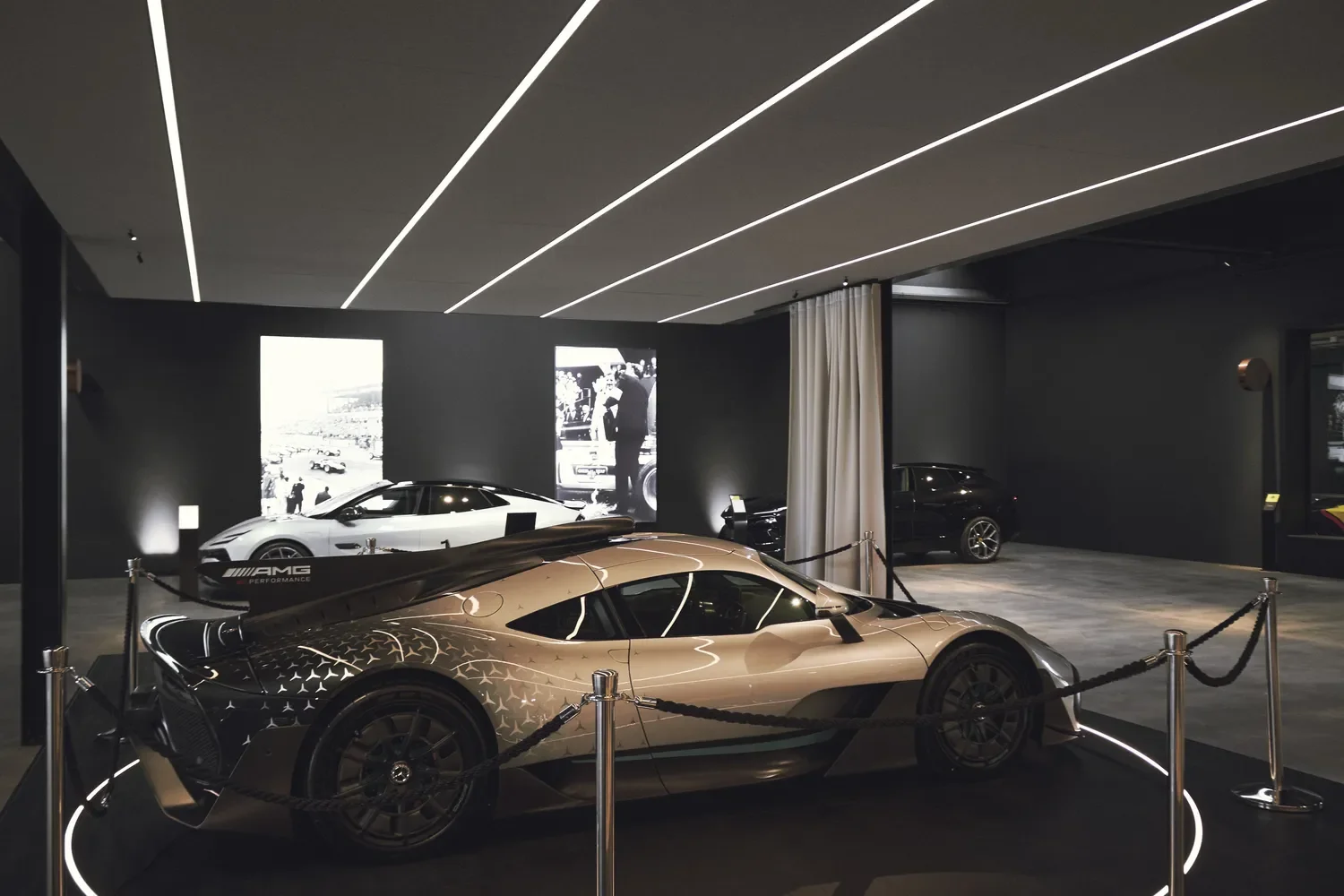 A luxury car exhibition featuring three high-end cars on display in a modern, dimly lit showroom with black walls and ceiling accent lighting. The central car is a beige supercar surrounded by a black rope barrier, with two other cars in the background, one white and one black, along with projected historical black-and-white photos on the wall.