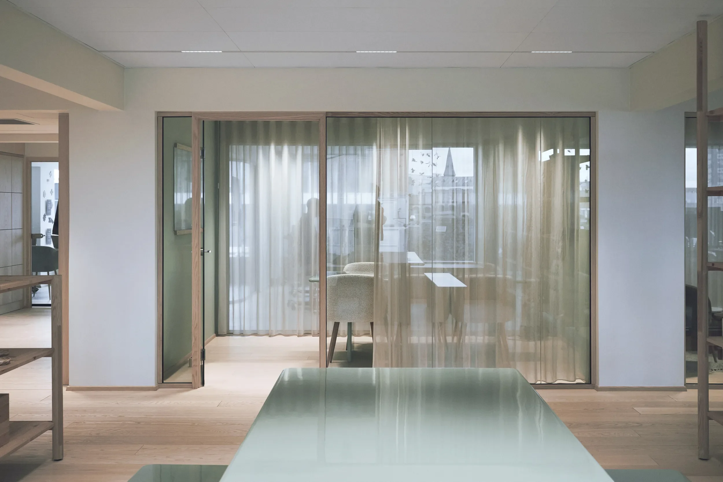 Modern office interior fand lighting design for Søstrene Grenes HQ with glass walls, curtains, a chair, and a view of rooftops and a church steeple outside.