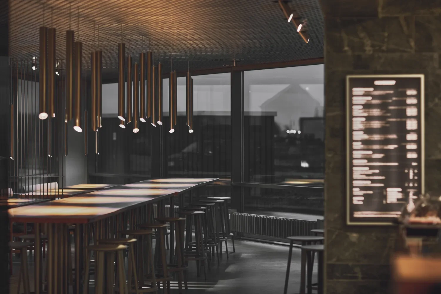 Empty modern restaurant interior with tall tables and bar stools, pendant lighting by LIK Lighting, large windows, and a blurred menu board on the right.