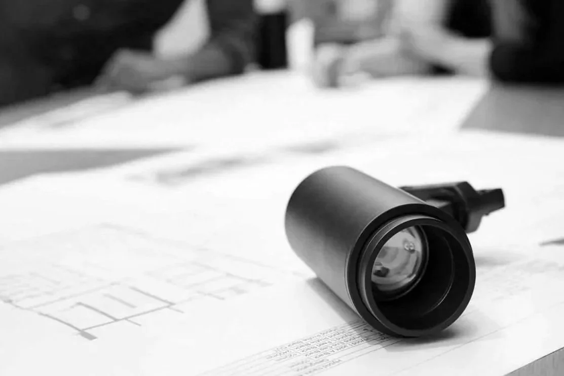 A black LIK Lighting spotlight resting on architectural plans on a table, with two people blurred in the background.