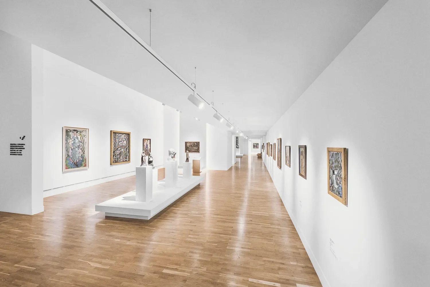Interior of an art gallery with framed paintings on white walls, sculptures on white pedestals, wooden flooring, and track lighting by LIK Lighting.