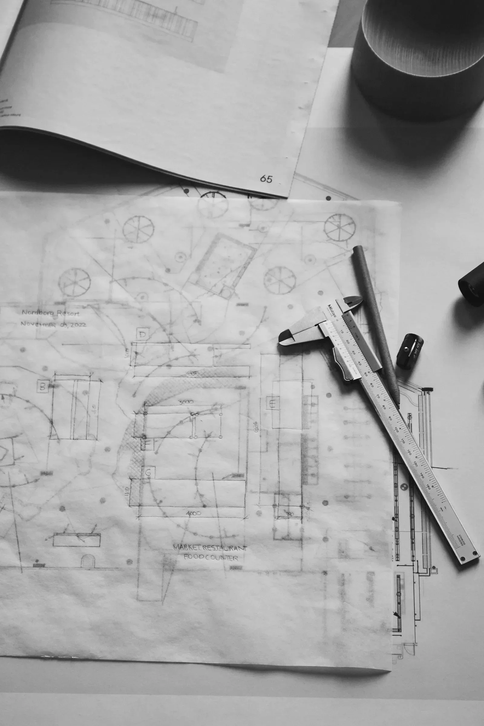 Engineering blueprint drawings spread out on a desk with a scale ruler, a pen, and a marker beside them, along with a cup on the top right corner.