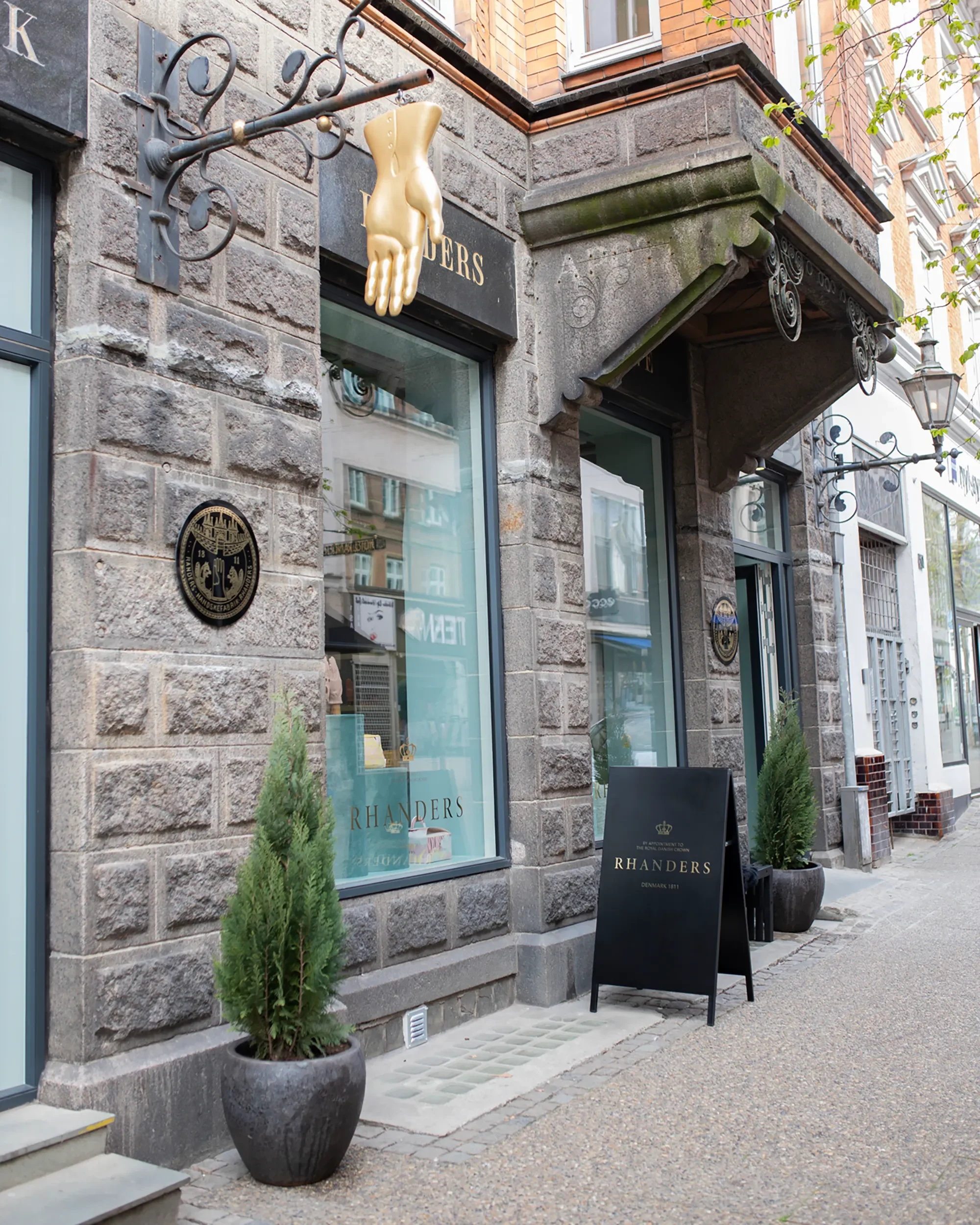 The image shows the exterior of Rhanders Handsker with stone walls, large windows, and decorative ironwork. There are potted plants, a black signboard & plaque on the wall. A gold-colored mannequin hand hangs from a wrought-iron bracket..