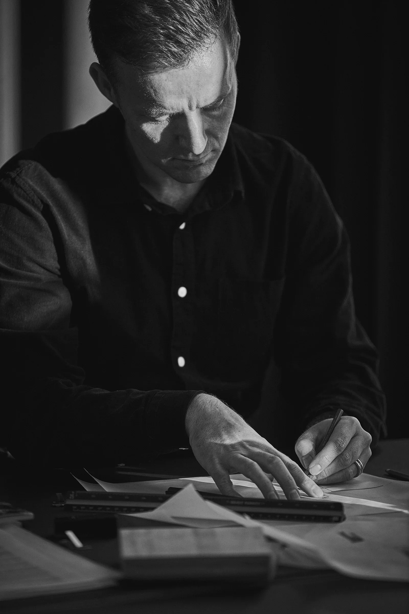 Black and white photo of LIK Lightings designer Lars Vejen writing on paper at a desk, with papers and files scattered around.