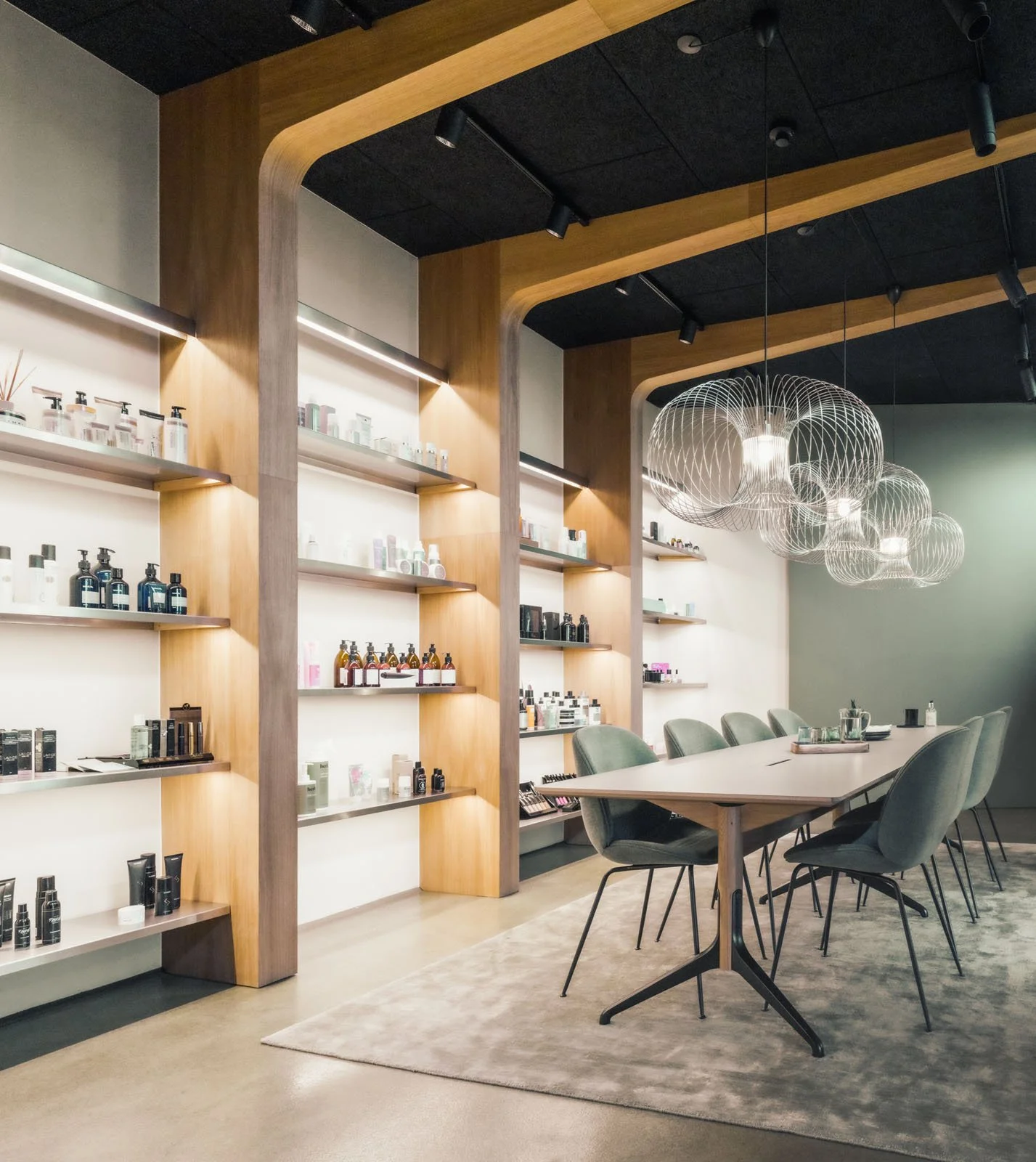Modern conference room with a long table, green chairs, and a wall with wooden shelves displaying bottles and products. Hanging spherical pendant lights and a neutral color palette.