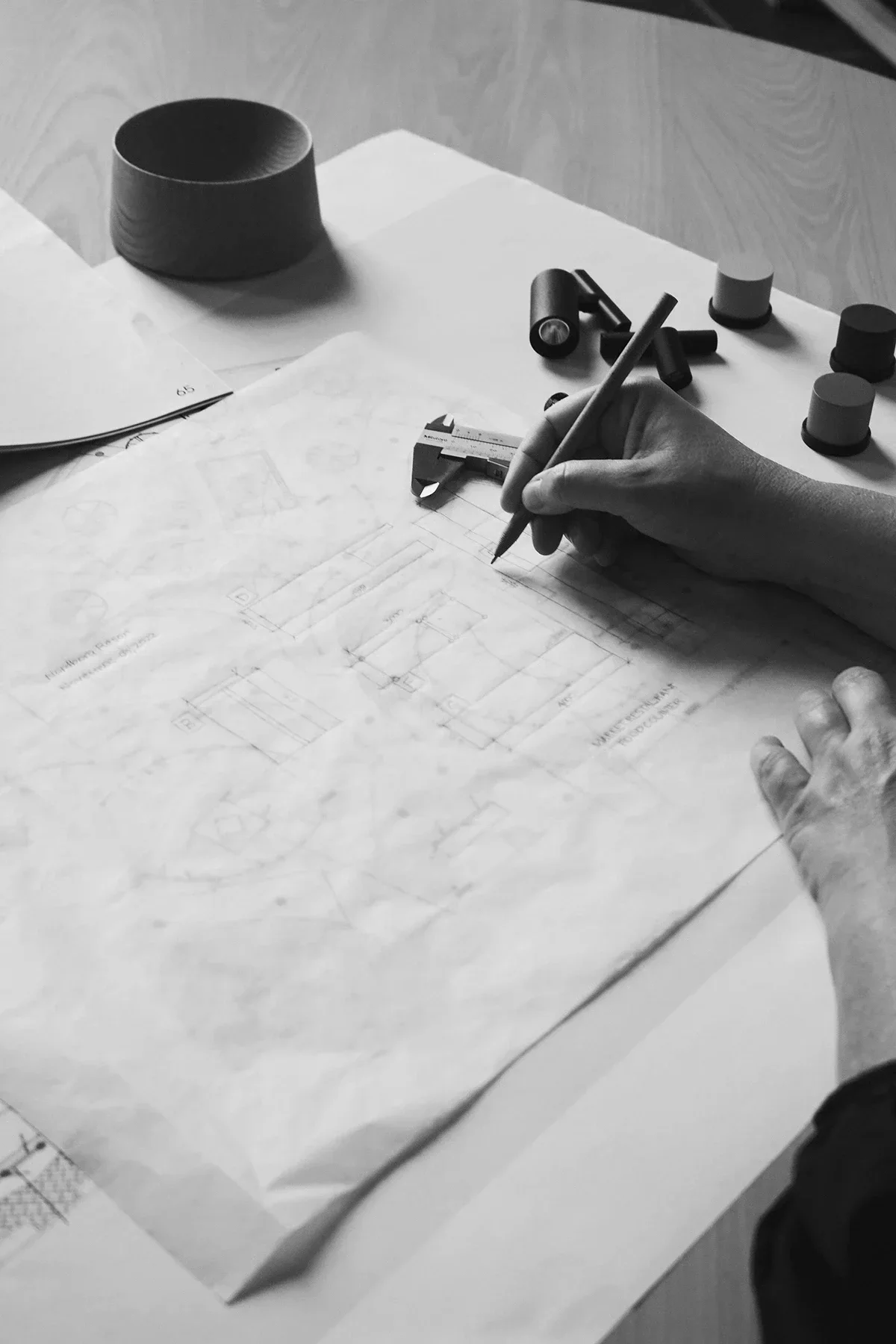 A lighting expert is working on architectural blueprints, using a caliper to measure. The workspace includes various small cylindrical objects and a large roll of tape, with papers and drawings spread out on a wooden table.