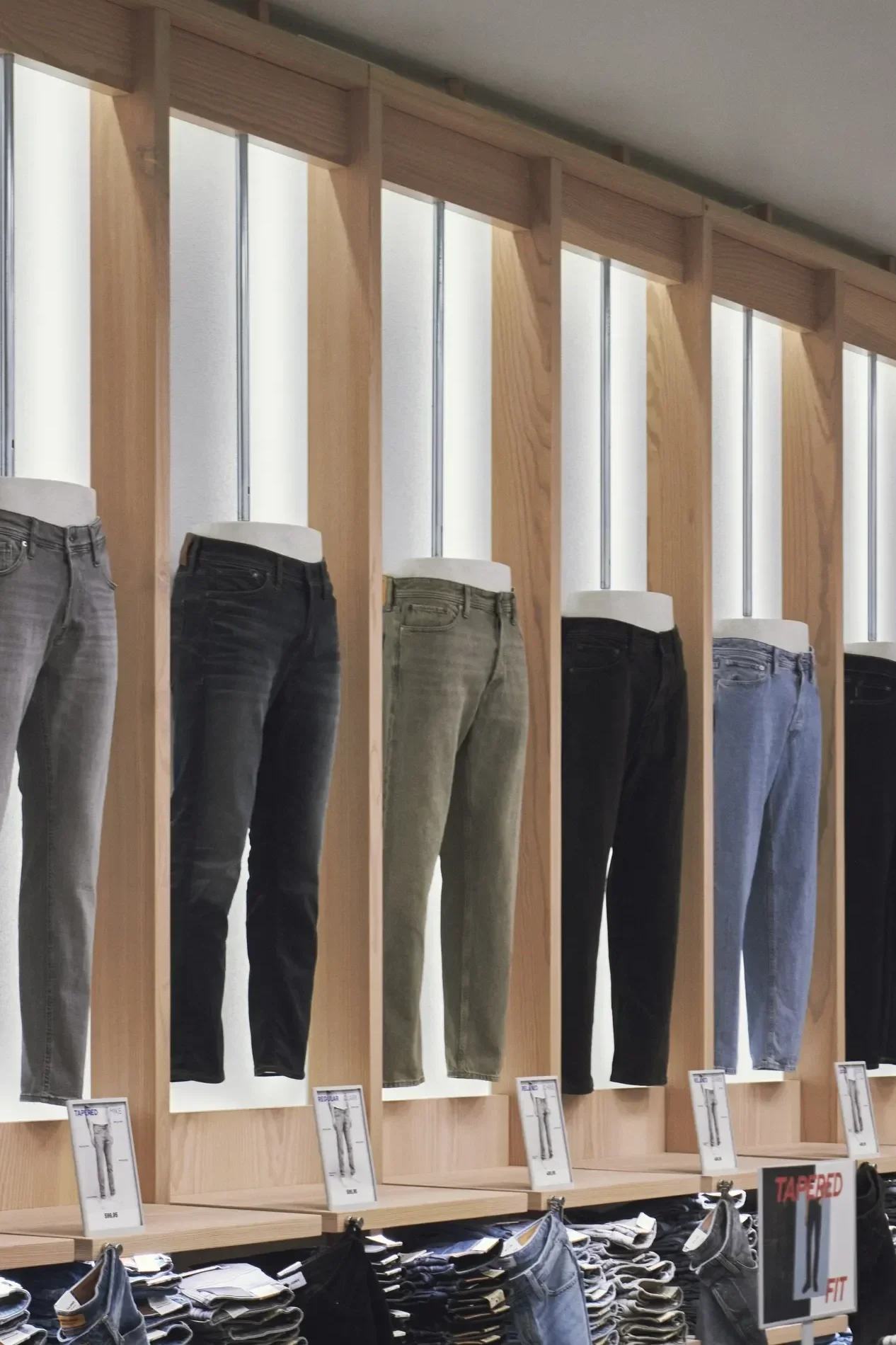 Display of jeans on mannequin legs at a clothing store, with different styles and washes, arranged vertically on wooden panels with lighting design.