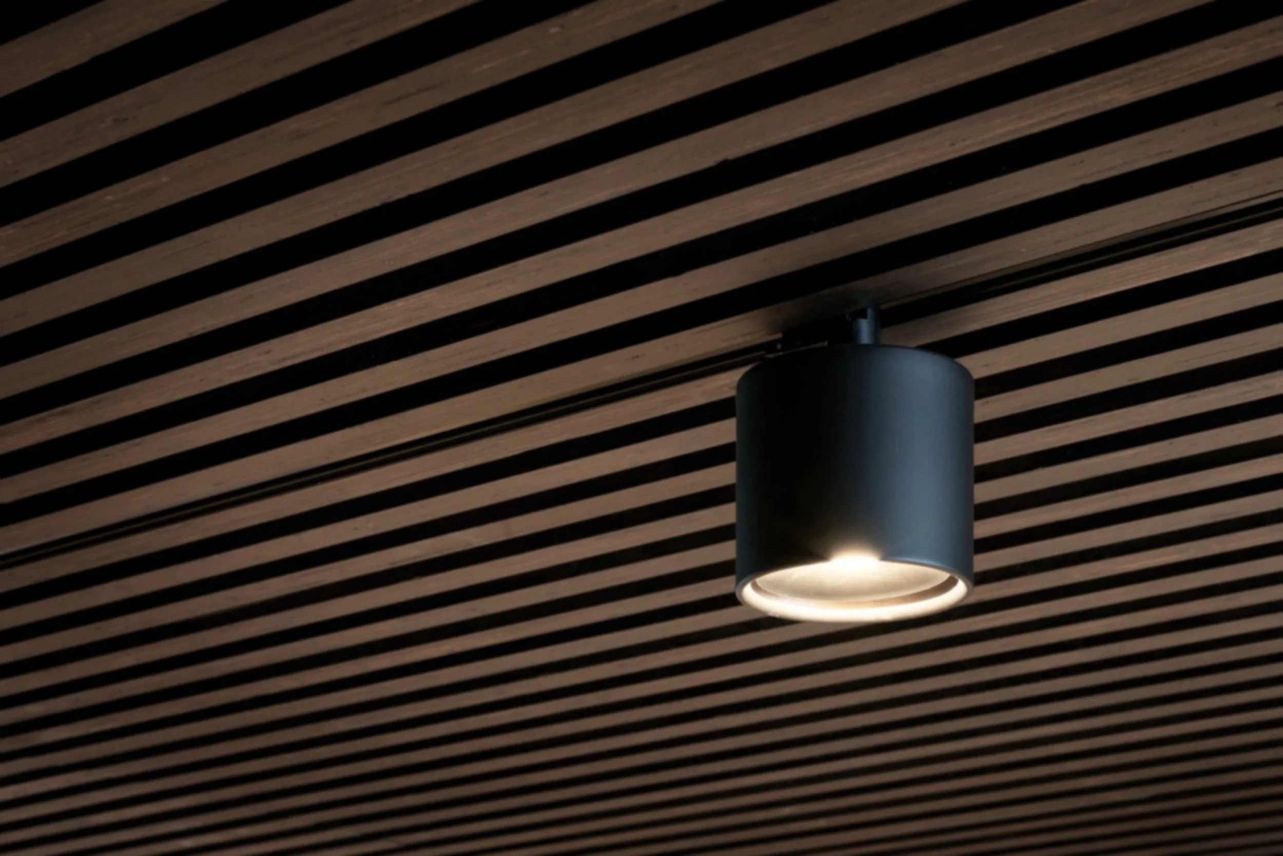 Recessed ceiling spotlight with wooden slats and a black cylindrical light fixture emitting a soft white light.