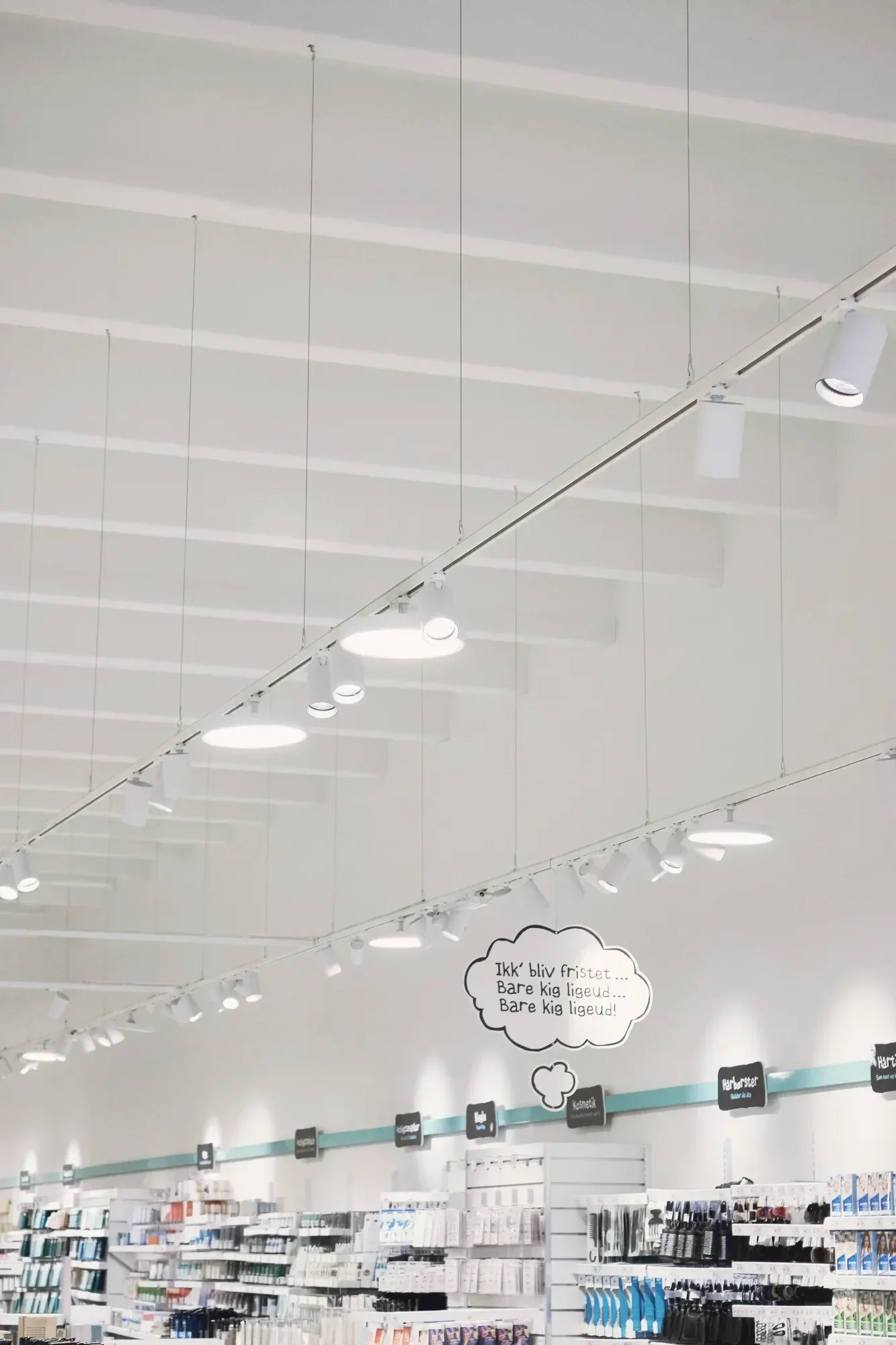 Bright Scandinavian pharmacy interior with suspended track lighting for NORMAL, product shelves, and Danish speech-bubble signage.
