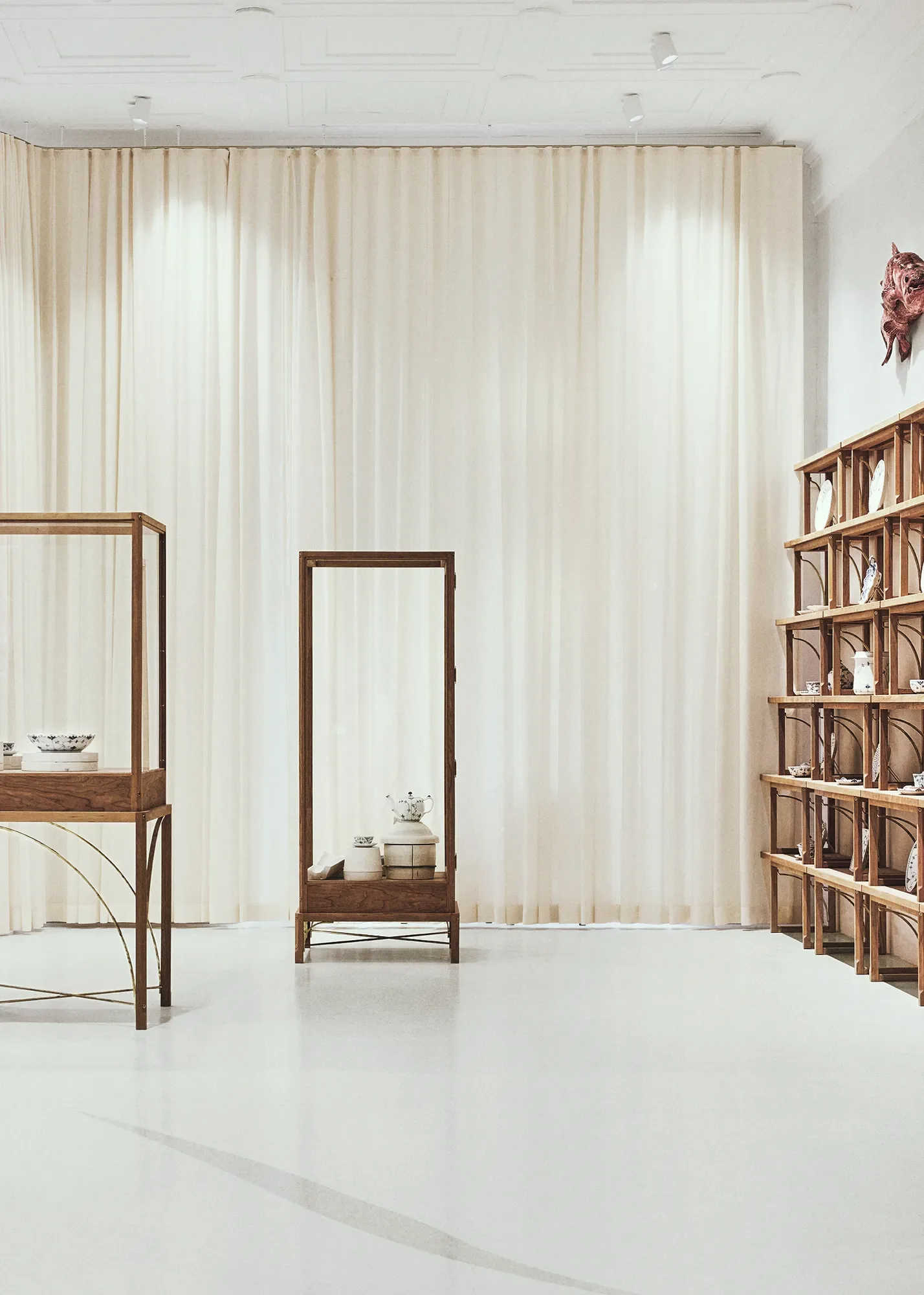 Display of lighting design by LIK Lighting for Royal Copenhagen such as cups and bowls arranged on wooden shelves and a glass cabinet, set against cream curtains in a minimalist room in Royal Copenhagen's Flagship Store.