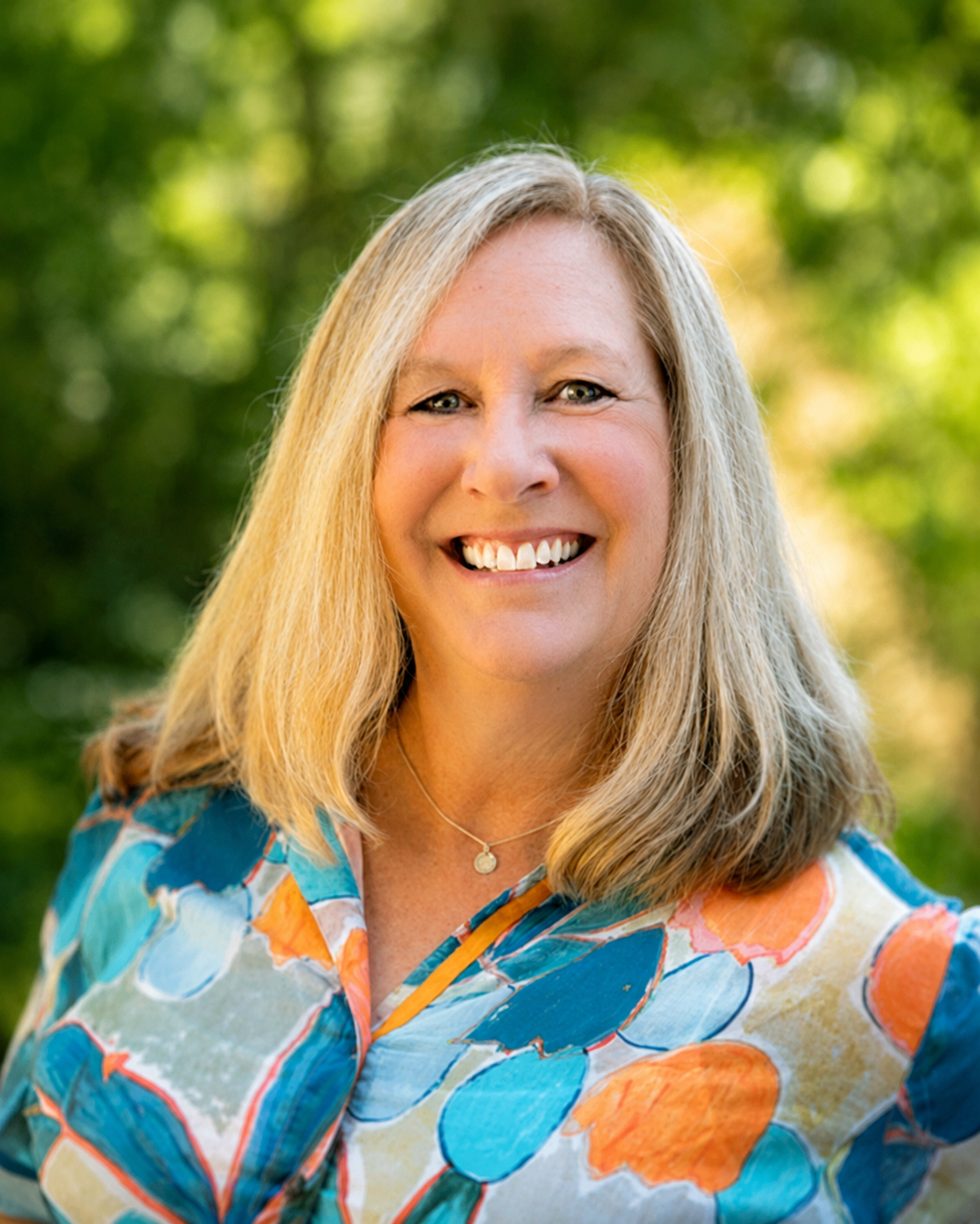A woman with blond hair smiling outdoors, wearing a colorful floral shirt with blue, orange, and yellow patterns, and a necklace, with green trees in the background.
