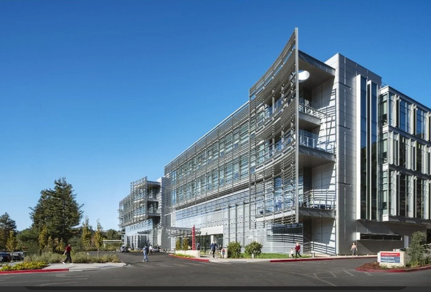 Modern multifloor office building with glass facade and aluminum sunshades, surrounded by a parking lot and trees, under a clear blue sky.