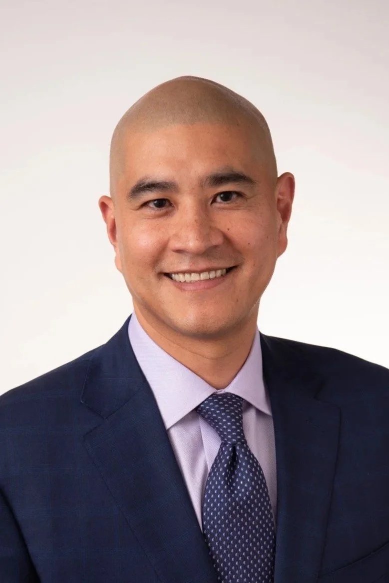 A professional headshot of a man in a dark suit, light purple shirt, and patterned tie, smiling against a plain white background.