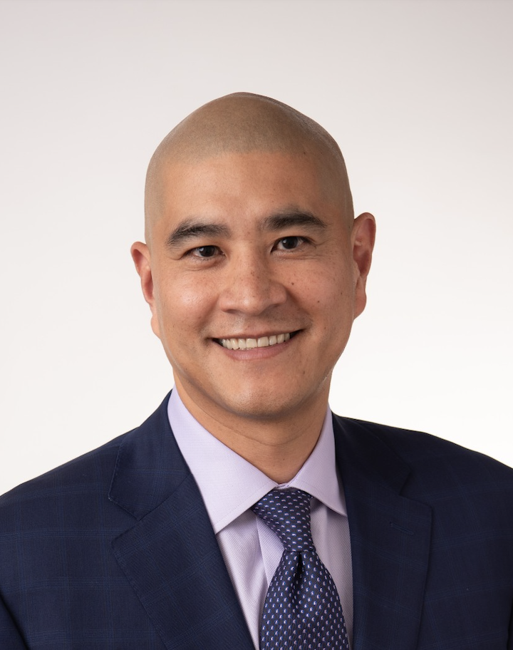 Professional headshot of an Asian man in a navy blue suit with a light purple shirt and patterned tie, smiling against a plain white background.