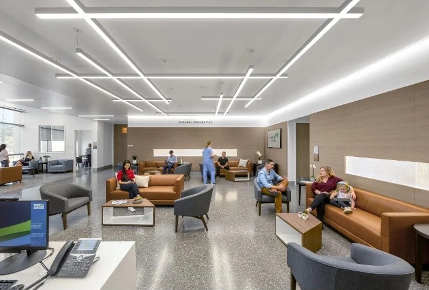 A hospital waiting room with modern furniture and people sitting. There are chairs, sofas, and a nurse walking through the room, some people using their phones, and a few waiting in the background.