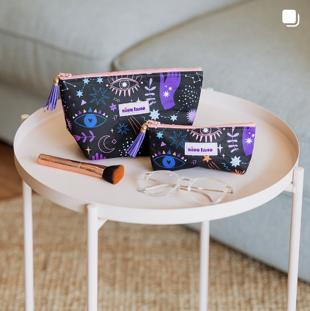 Two cosmetic bags with eye and moon designs for Sisu Lane, a makeup brush, and a pair of eyeglasses on a round white table.