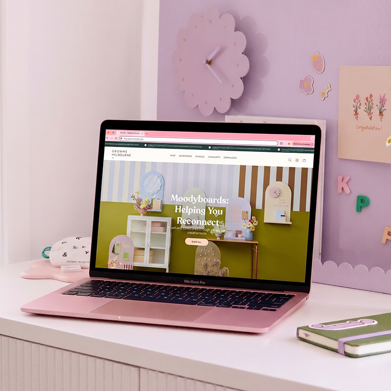 A pink MacBook Pro on a white desk, displaying a webpage for GROWME Melbourne with colorful indoor decor, and behind it a purple wall with a cloud-shaped clock and decorative stickers of pastel-colored strawberries, flowers, and letters.