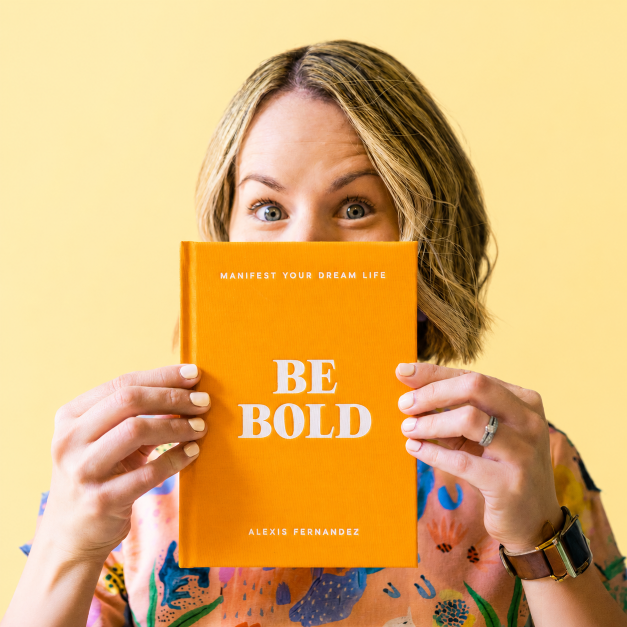 A woman holding an orange book titled "Be Bold" in front of her face, with wide eyes visible above the book. The book's subtitle reads "Manifest Your Dream Life" and it's authored by Alexis Fernandez. The woman is wearing a colorful, patterned shirt, a wristwatch, and a ring, with a solid yellow background behind her.