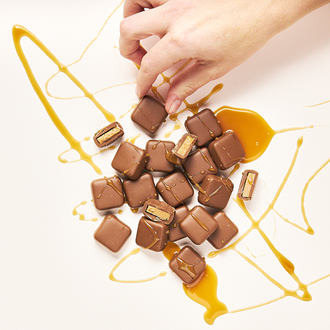 Hand picking up a Coated chocolate-covered caramels from a scattered pile of similar chocolates on a white surface, with caramel drizzled around them.