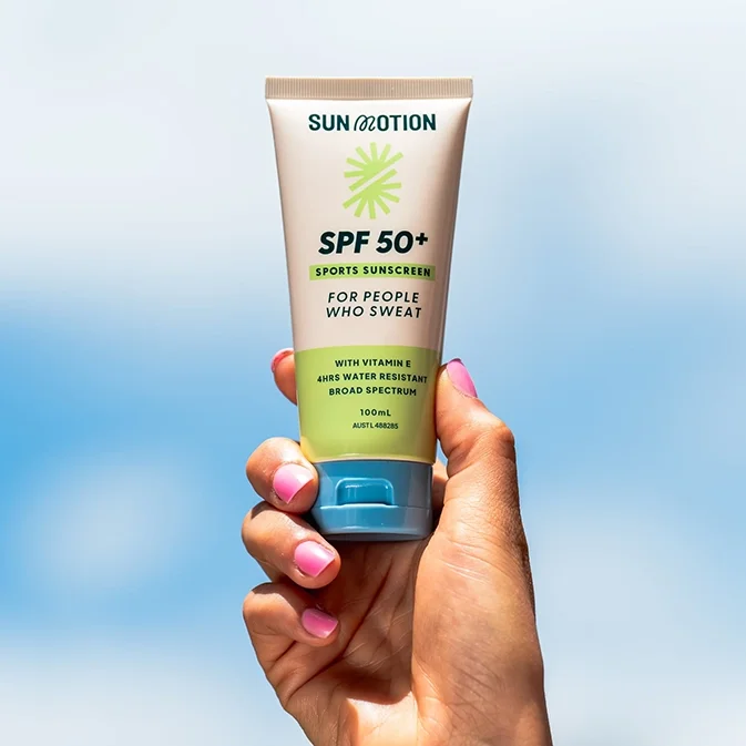 A hand holding a tube of Sun Motion SPF 50+ sports sunscreen against a blue sky background.