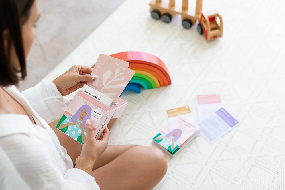 A person sitting on the floor sorting a colorrful card deck called Emotional Support for Mothers with a rainbow wooden toy in the background.
