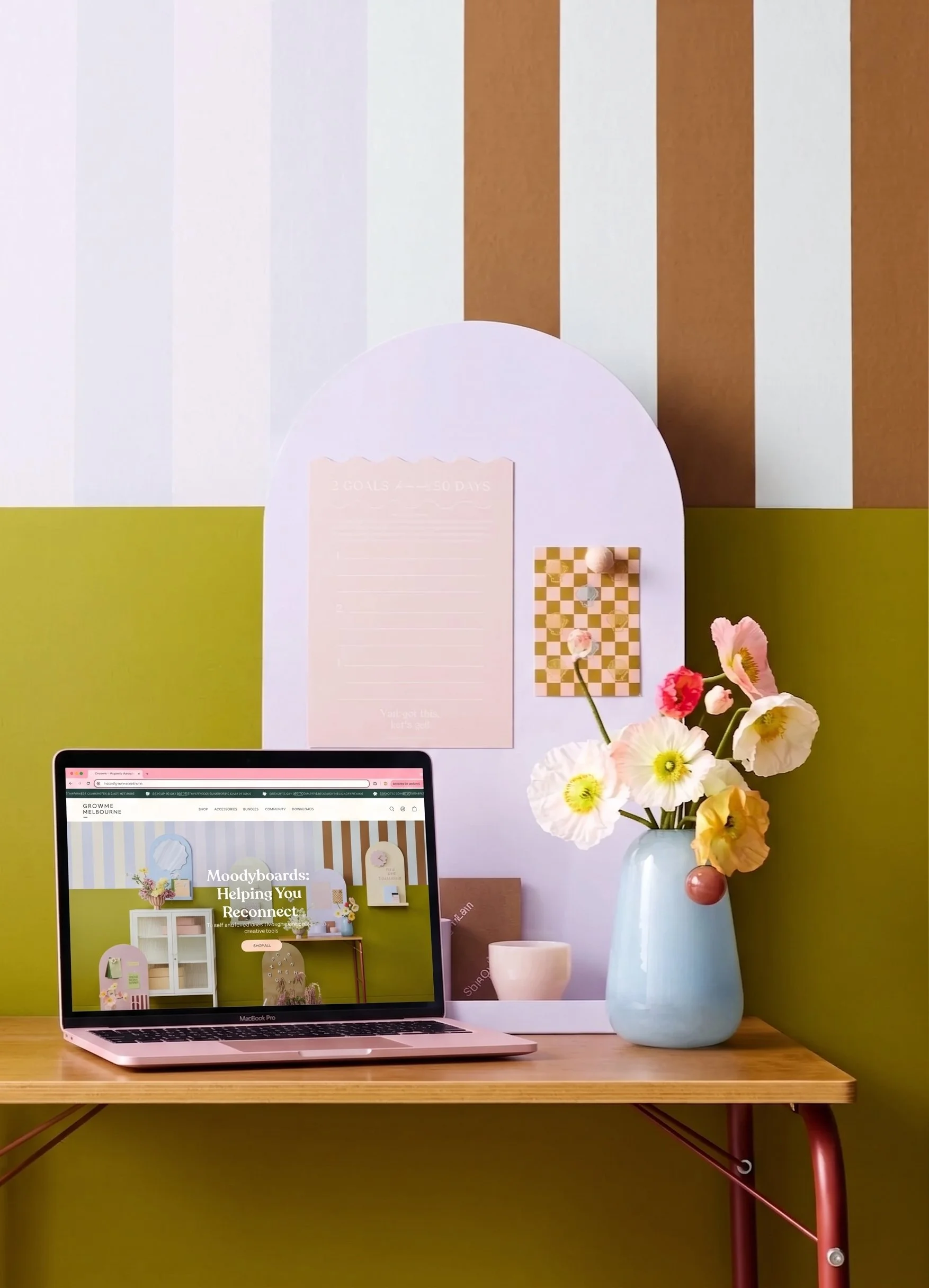 A workspace with a pink laptop on a wooden desk, a blue vase with colorful flowers, a beige cup, and a background featuring striped wall decor and a larger screen displaying a website outline.