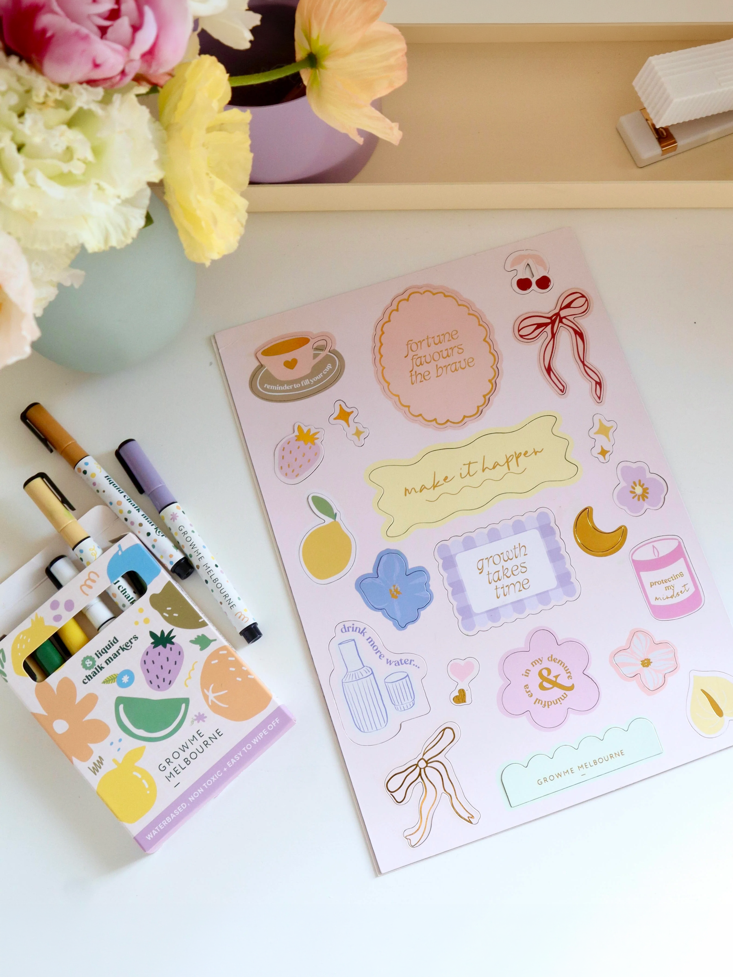 Colorful stationery and decorative stickers on a white desk with a bouquet of pink, yellow, and white flowers in a pale green vase.