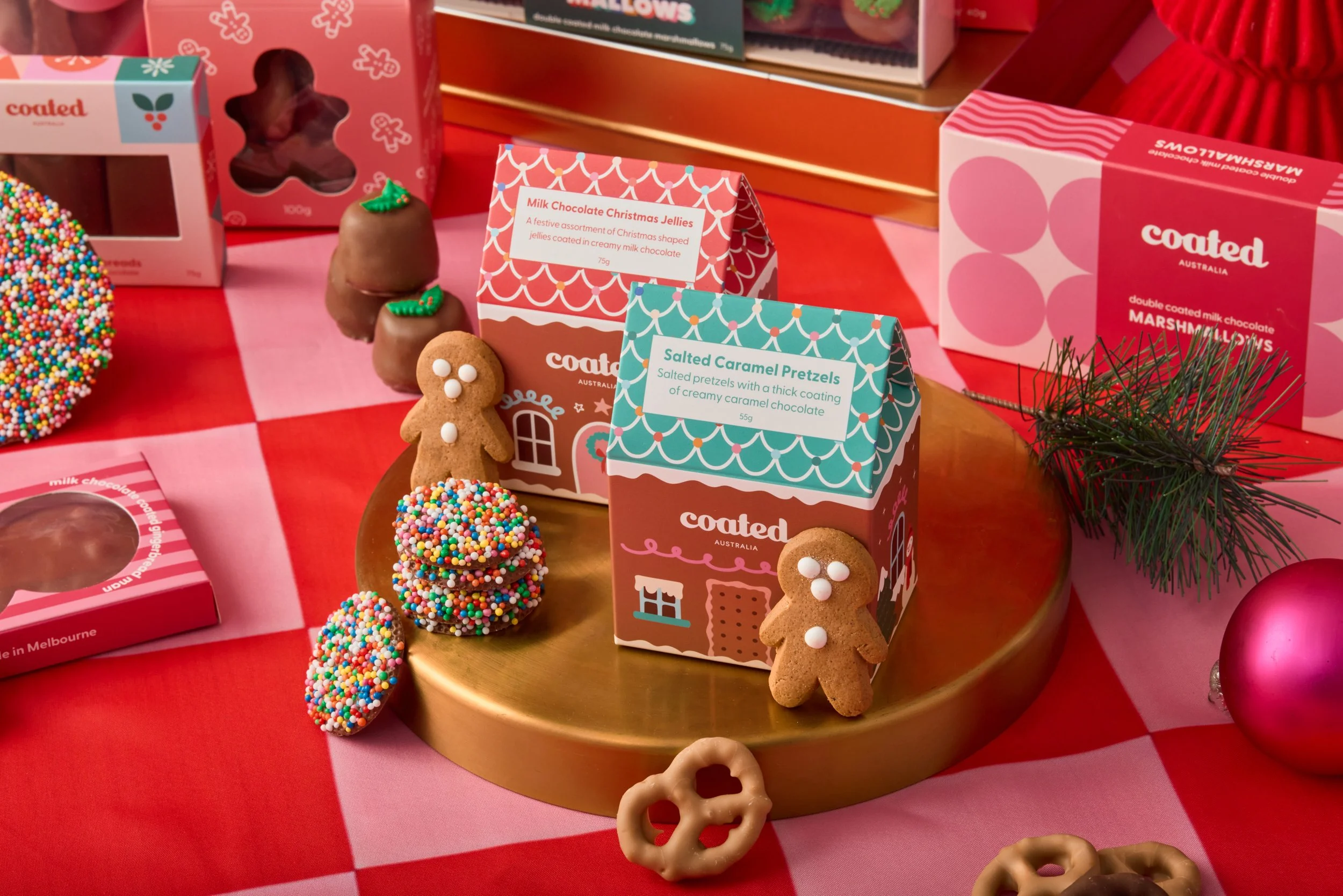 Display of holiday-themed chocolates and treats, including gingerbread cookies, pretzels, and assorted chocolates in colourful Coated packaging on a red and pink checkered tablecloth, with some greenery and ornaments in the background.