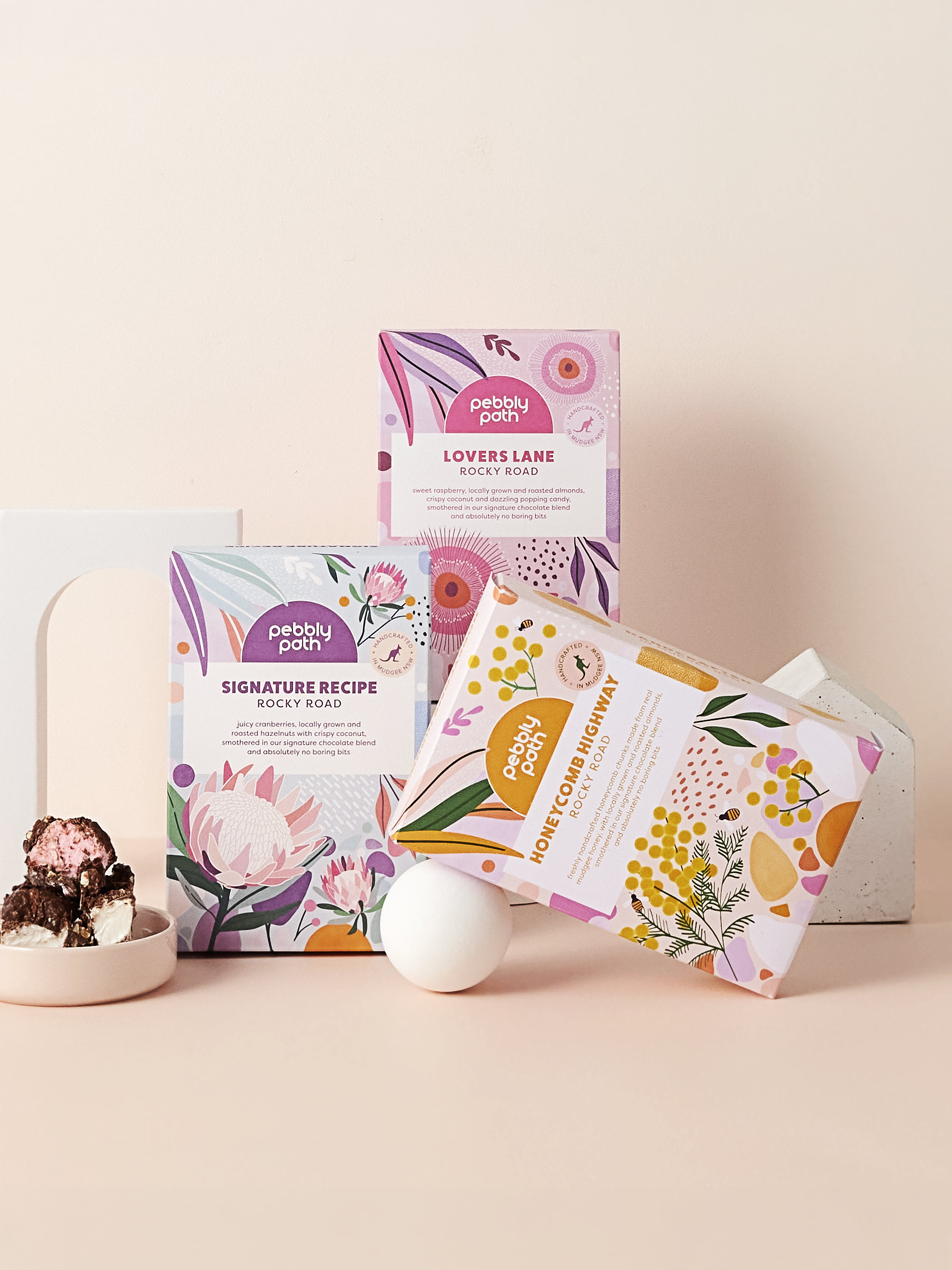 Colorful boxes of Pebbly Path chocolates with floral and abstract designs, a bowl of chocolate chunks with marshmallows, and white eggs on a pink background.