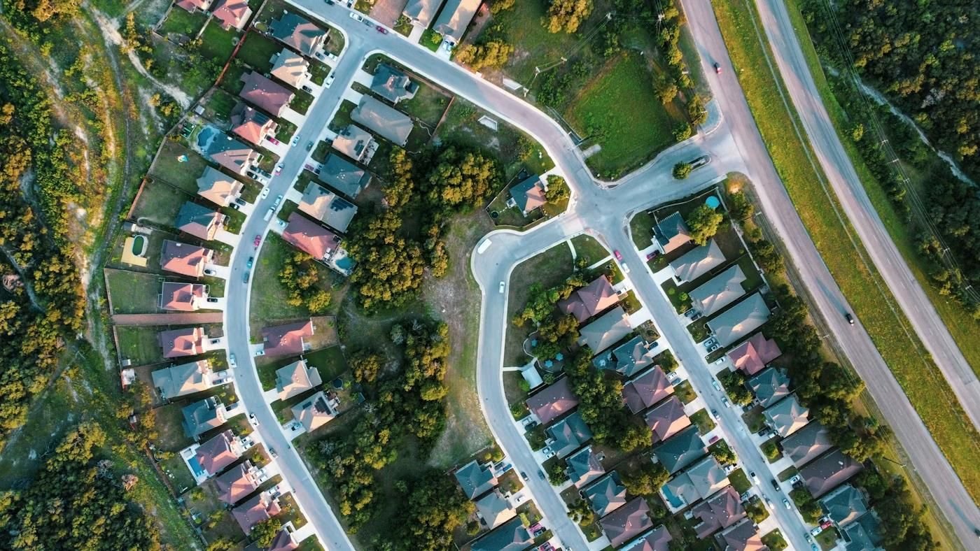 Aerial view of a suburban neighborhood with winding streets, houses with driveways, green lawns, trees, and a nearby highway.