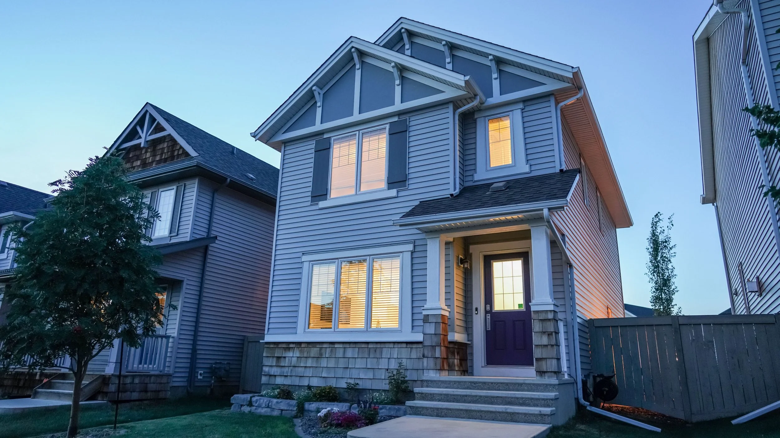 A multi-story house with gray siding, illuminated windows, and a front porch, surrounded by a small yard with a tree and plants, during dusk.