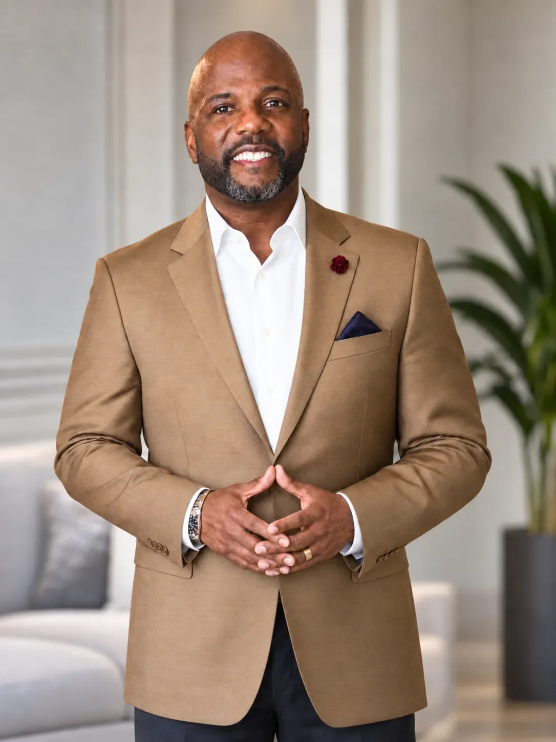 A man with a beard and bald head, wearing a beige blazer with a white shirt, stands inside a modern, well-lit room with gray walls and a large green plant in the background.