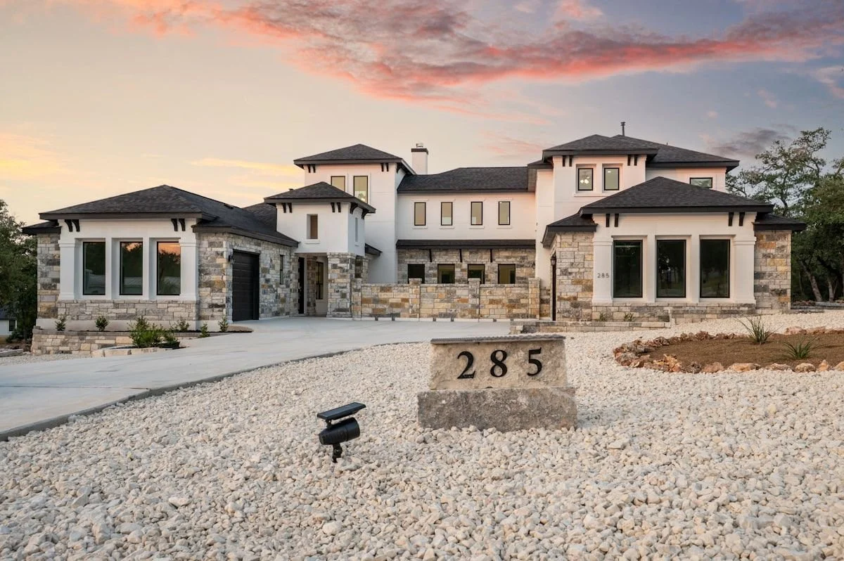 Exterior of a large, modern house with stone and white walls, multiple windows, and a black roof, during sunset with pink clouds in the sky. A concrete driveway leads up to the house, and a stone marker displays the house number 285. Landscaping includes gravel and small plants.