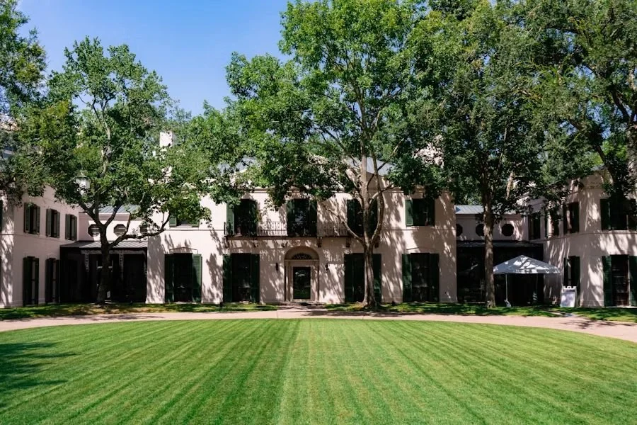 A large beige building with black window shutters, surrounded by tall green trees and a neatly mowed green lawn under a clear blue sky.