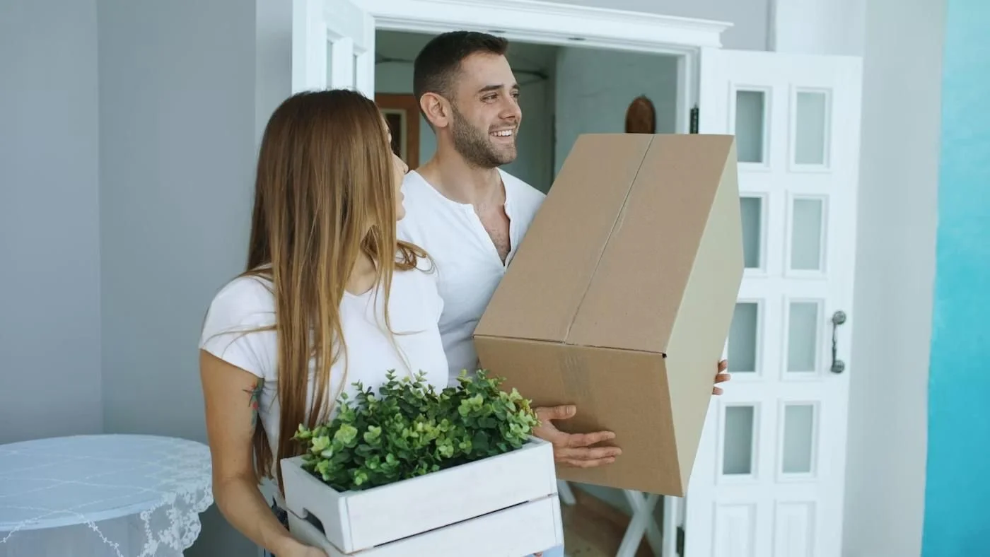 A man and woman smiling inside a house, holding boxes with plants, possibly moving in or out.