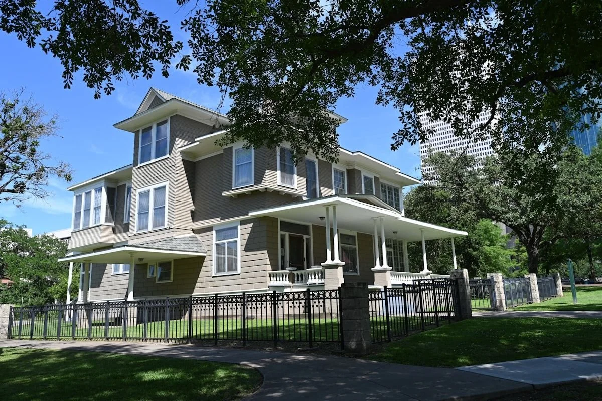 A large, three-story Victorian-style house with grey siding, a wrap-around porch, fenced yard, and multiple windows, set against a clear blue sky with trees surrounding it.