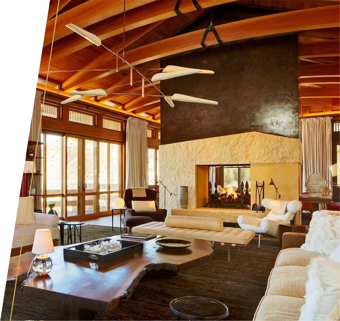 A cozy living room with large windows, a fireplace, and modern furniture, featuring wooden ceiling beams and warm lighting.