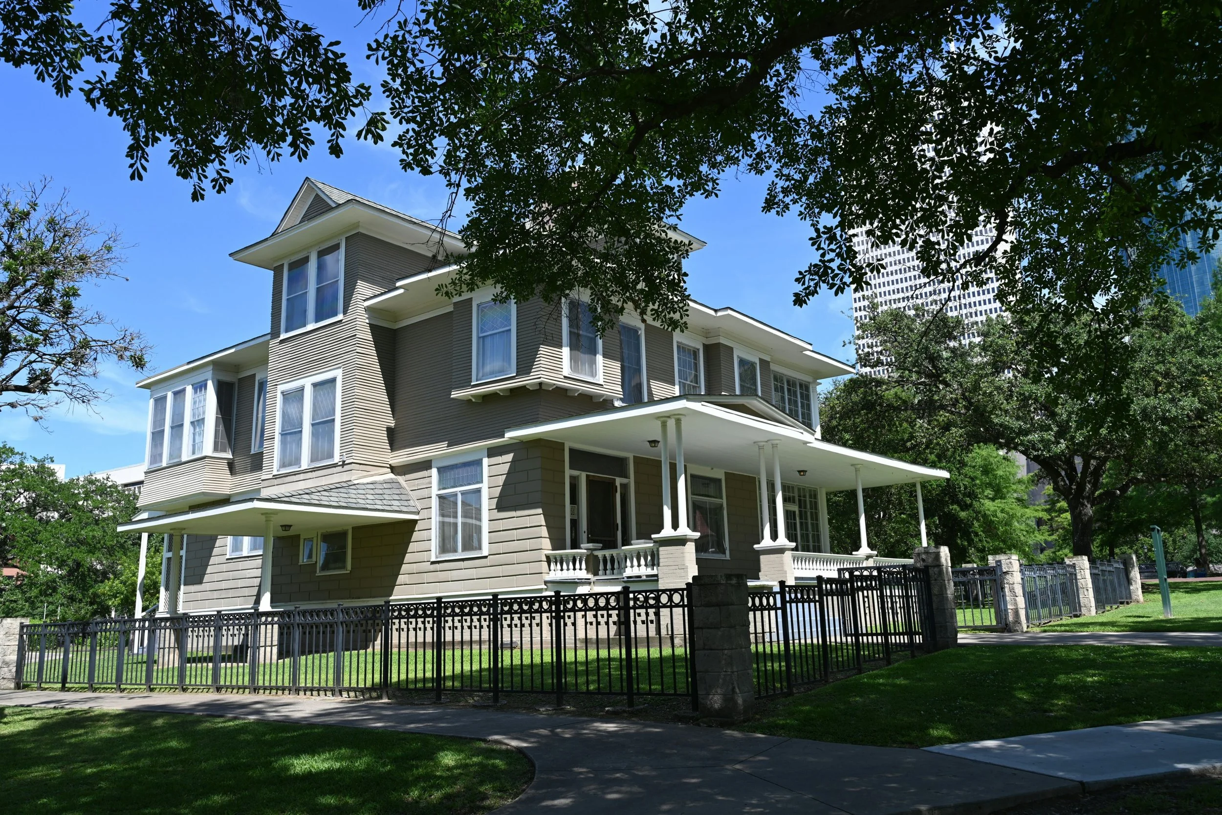 A large, multi-story house with beige siding and white trim, surrounded by green trees and lawn, under a clear blue sky.