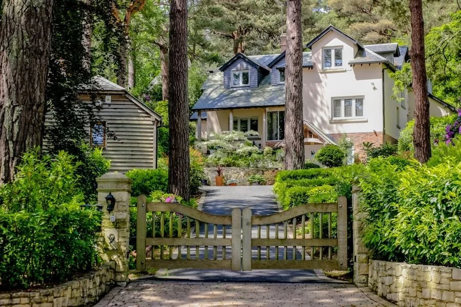 A house surrounded by tall trees and lush green bushes, with a wooden gate at the front and a stone pathway leading to the front porch.