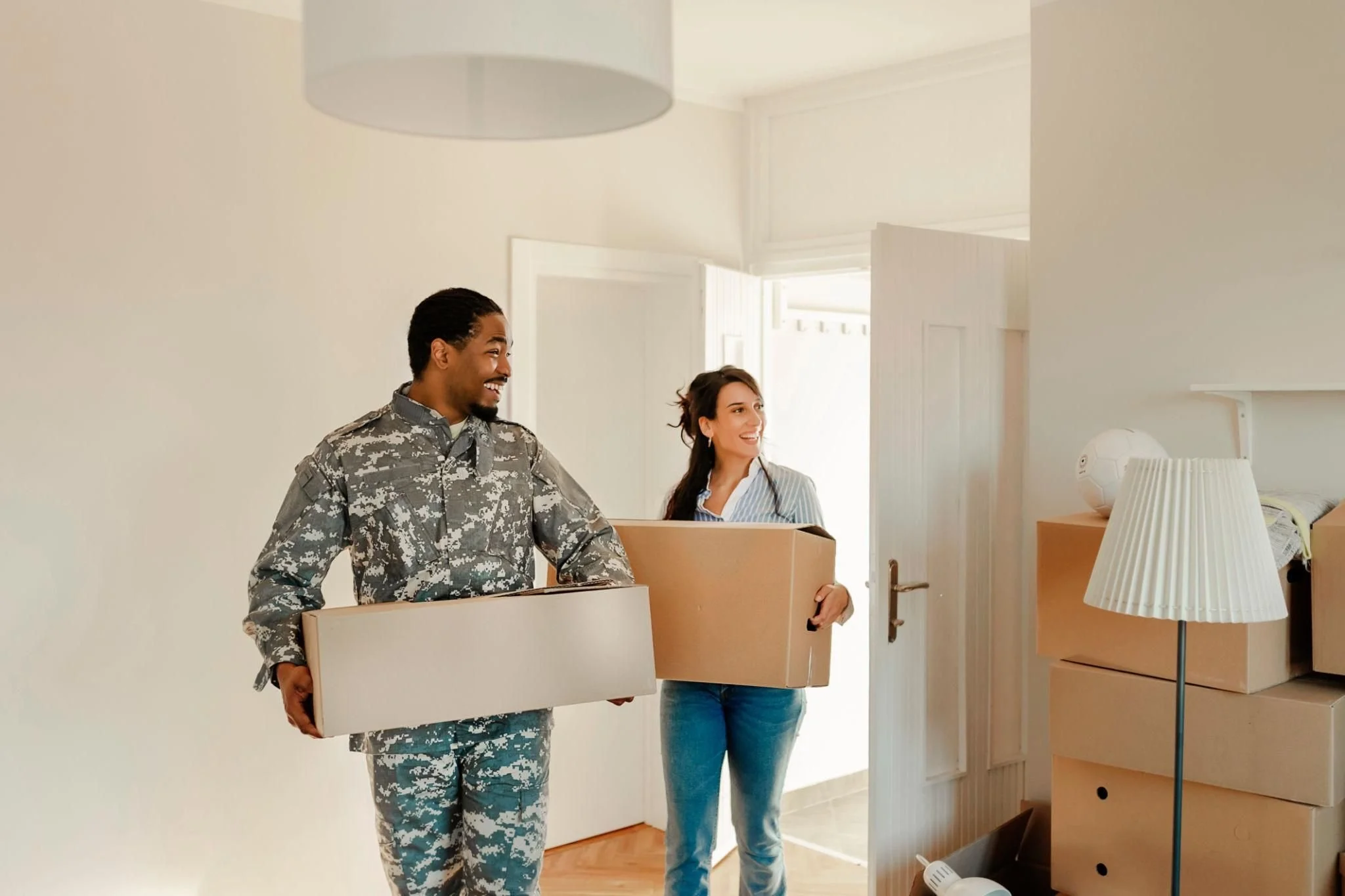 Two people, a man in military camouflage uniform and a woman in a striped shirt, carrying cardboard boxes into a room, smiling and happy.