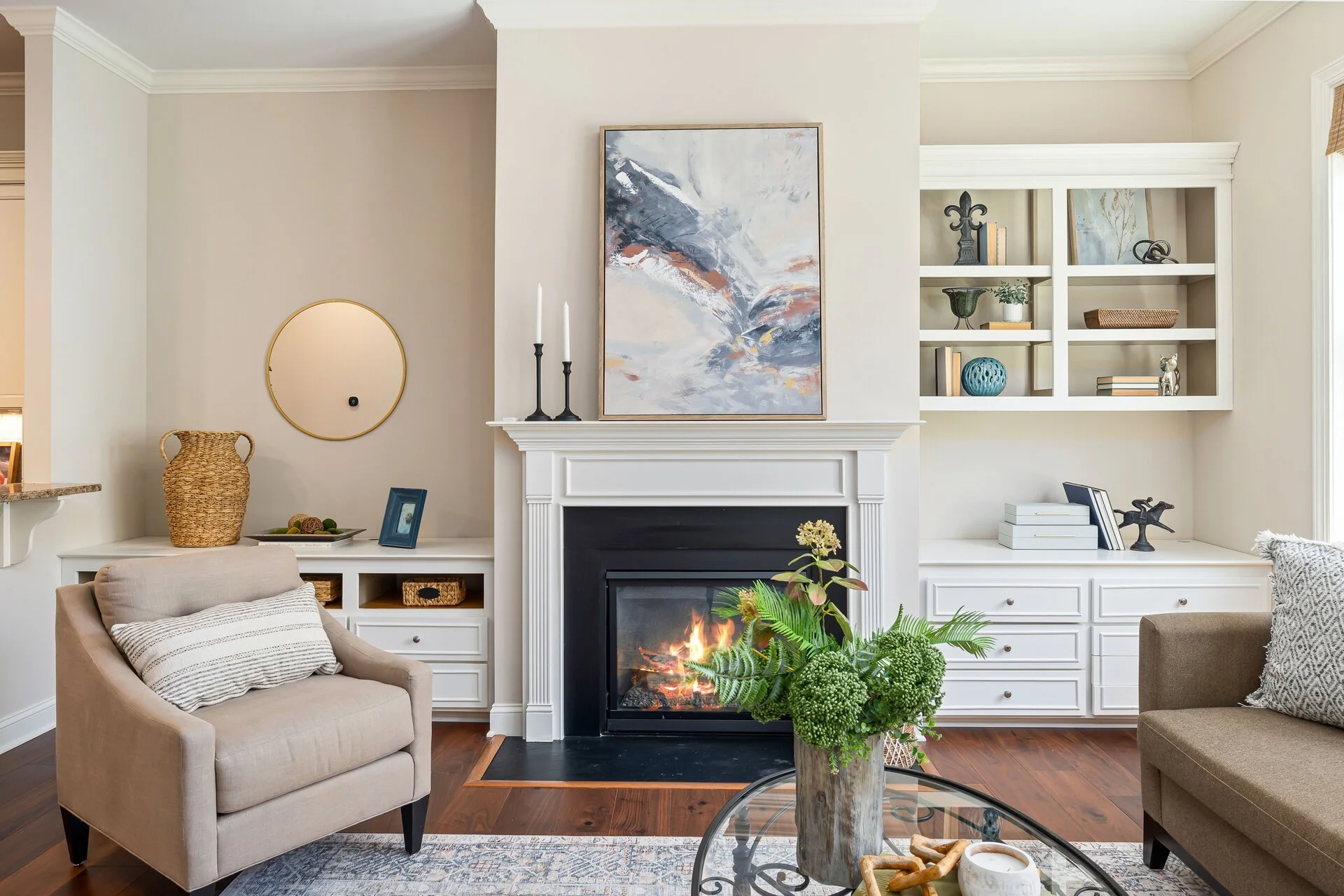 Living room with a beige armchair with striped pillow, a fireplace with a painting above it, and built-in white shelves with decorative items, adjacent to a window with blinds.