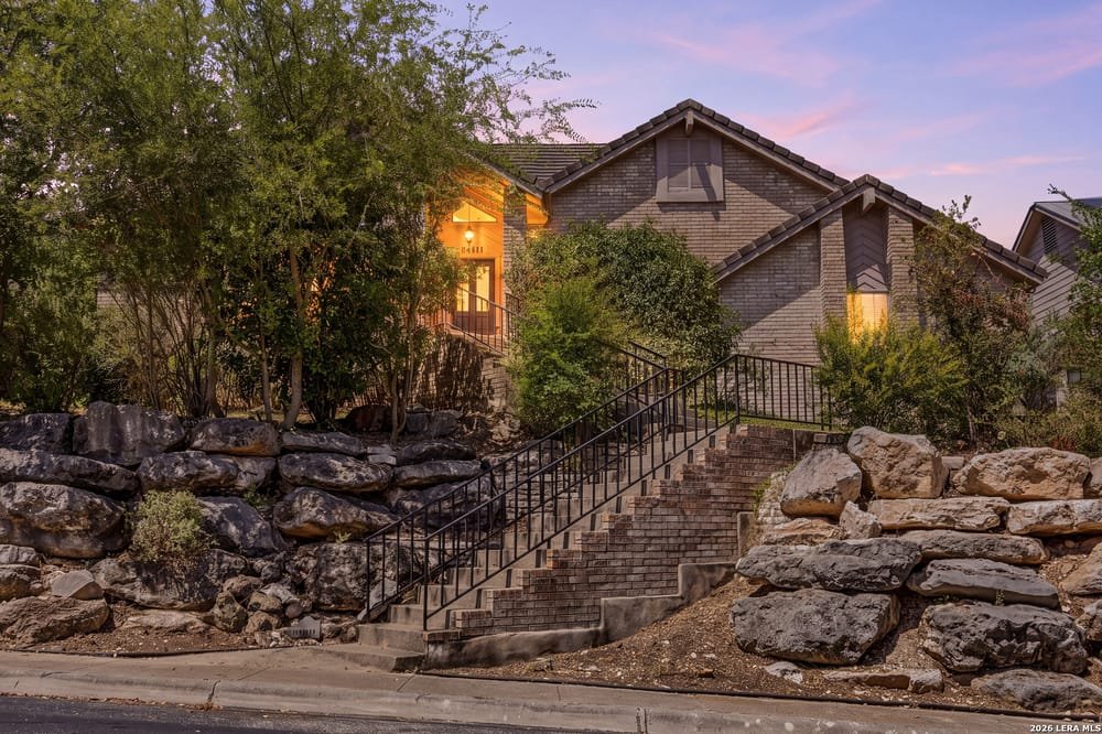 Residential house with brick exterior, set on a hill with a staircase leading up, surrounded by trees and rocks, during dusk.