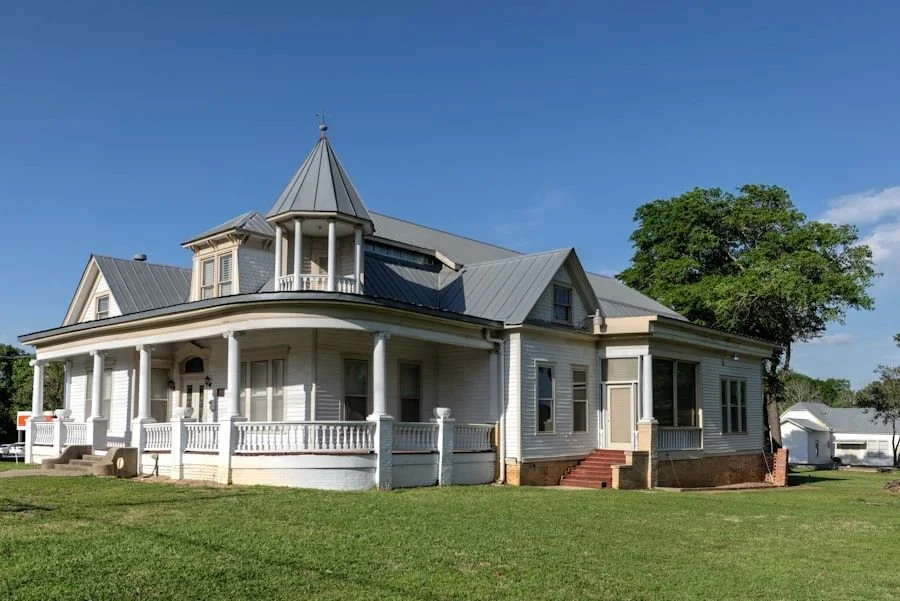 Large, historic white house with Victorian architectural features, a circular turret, wraparound porch, and a grassy lawn under a clear blue sky.