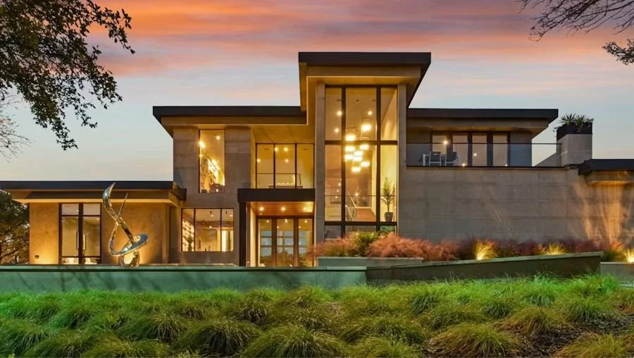 Modern house with large glass windows and a landscaped front yard, illuminated at sunset.