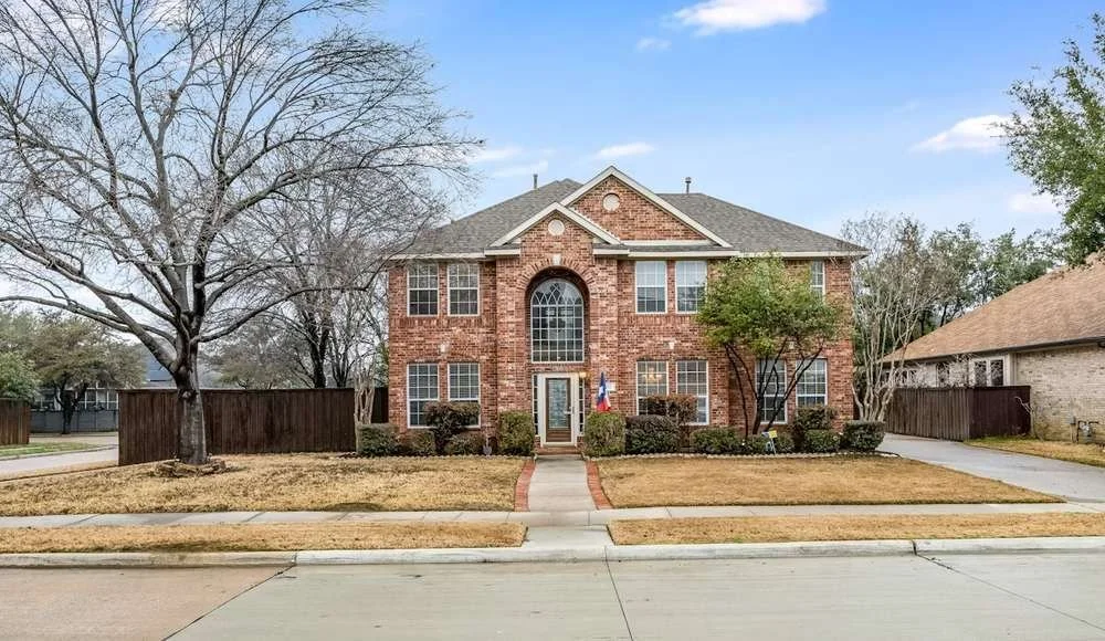 A two-story brick house with a gray roof, surrounded by a well-maintained lawn and trees, with a sidewalk in front and a driveway to the right.