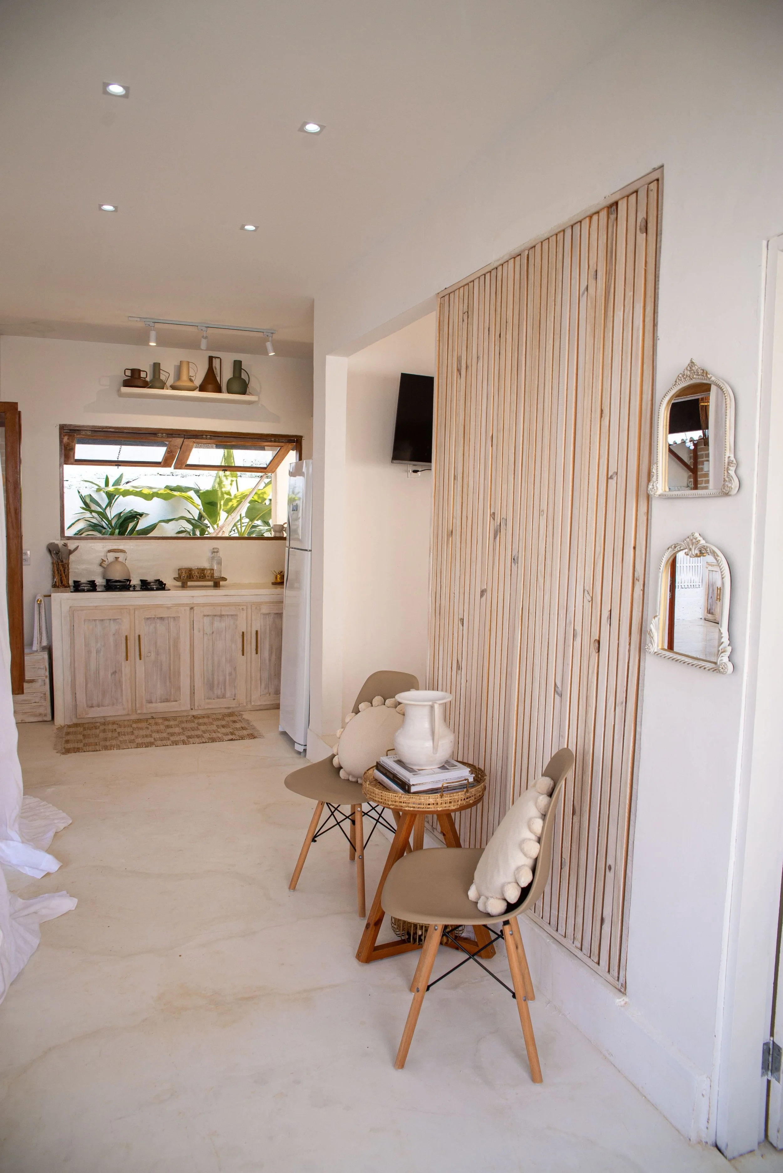 A cozy living space featuring a small table with two beige cushioned chairs, one adorned with a white pillow with pom-pom trim. The room has a light, neutral color palette with white walls and a light wooden slatted divider. A mirror with an ornate white frame hangs on the wall, and a flat-screen TV is mounted nearby. In the background, a kitchen area with wooden cabinets, a window with plants outside, and a shelf with decorative vases.
