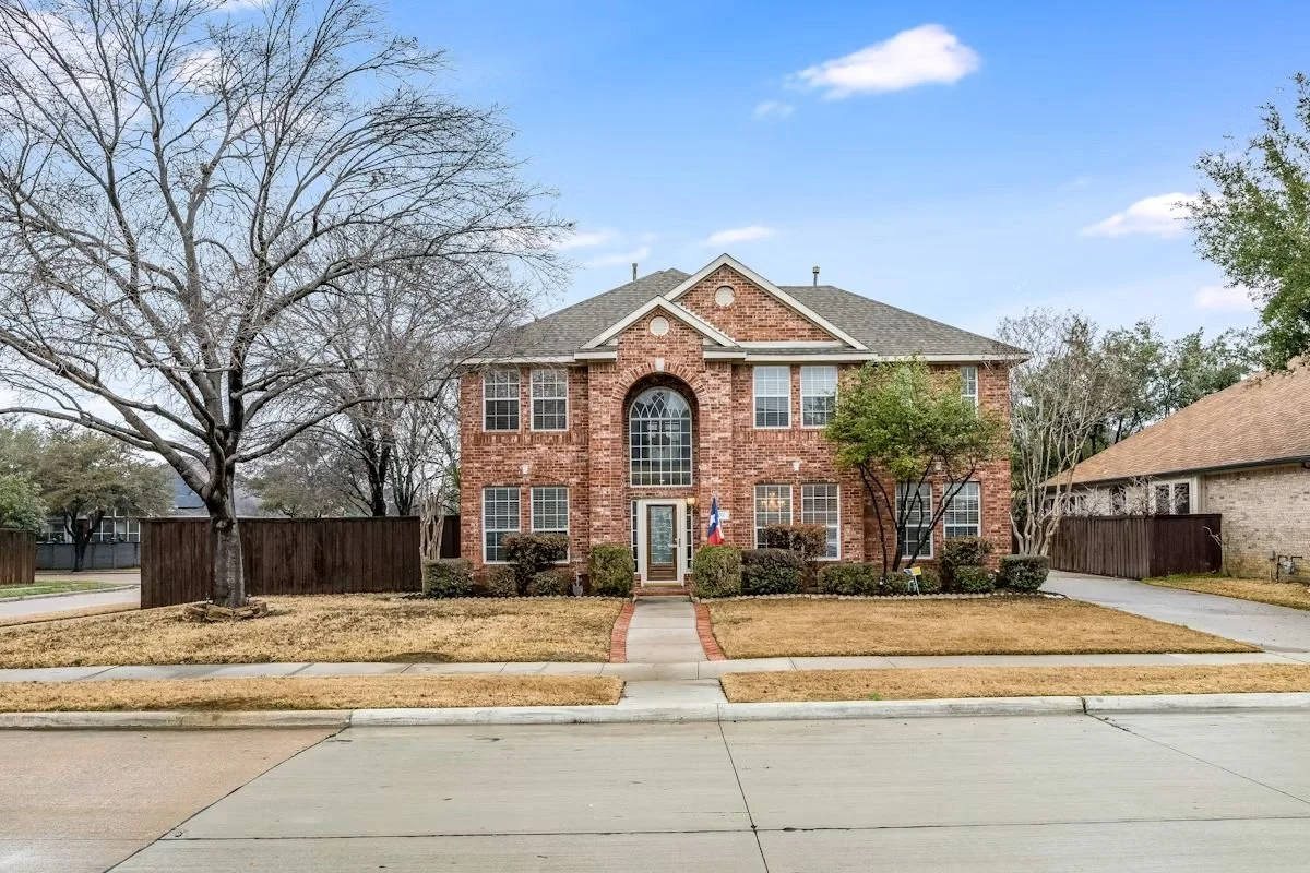 A two-story brick house with a gray roof, four front windows, a glass front door, and two trees in the front yard. The lawn is brown and dry, and there is a sidewalk in front.