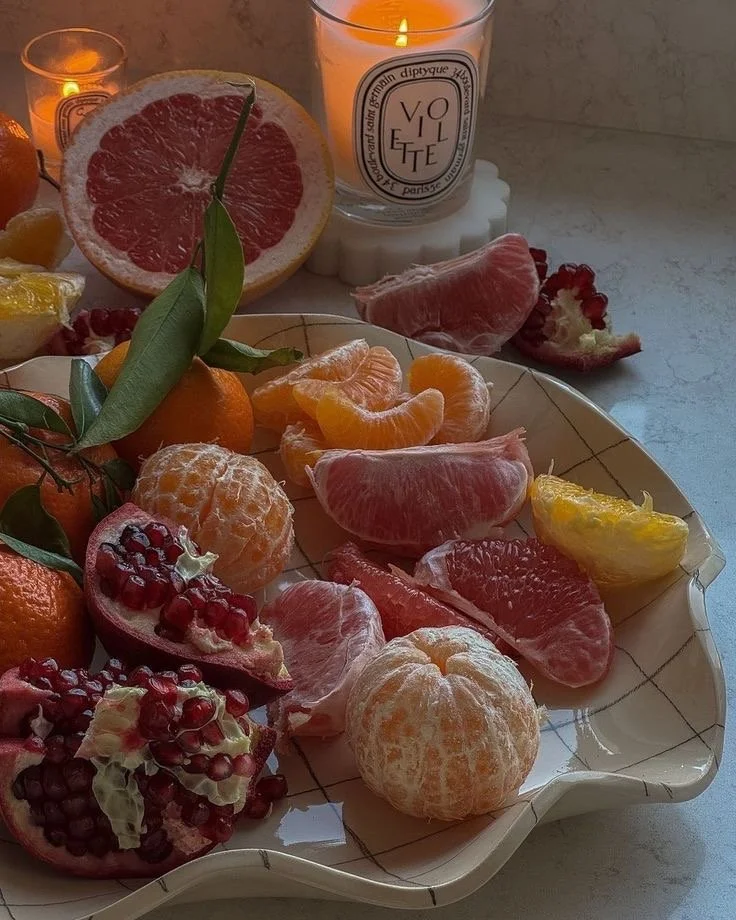 A decorative plate filled with various peeled and unpeeled citrus fruits, including oranges, tangerines, pomegranate, and grapefruit. In the background, there are candles and a glass of water.