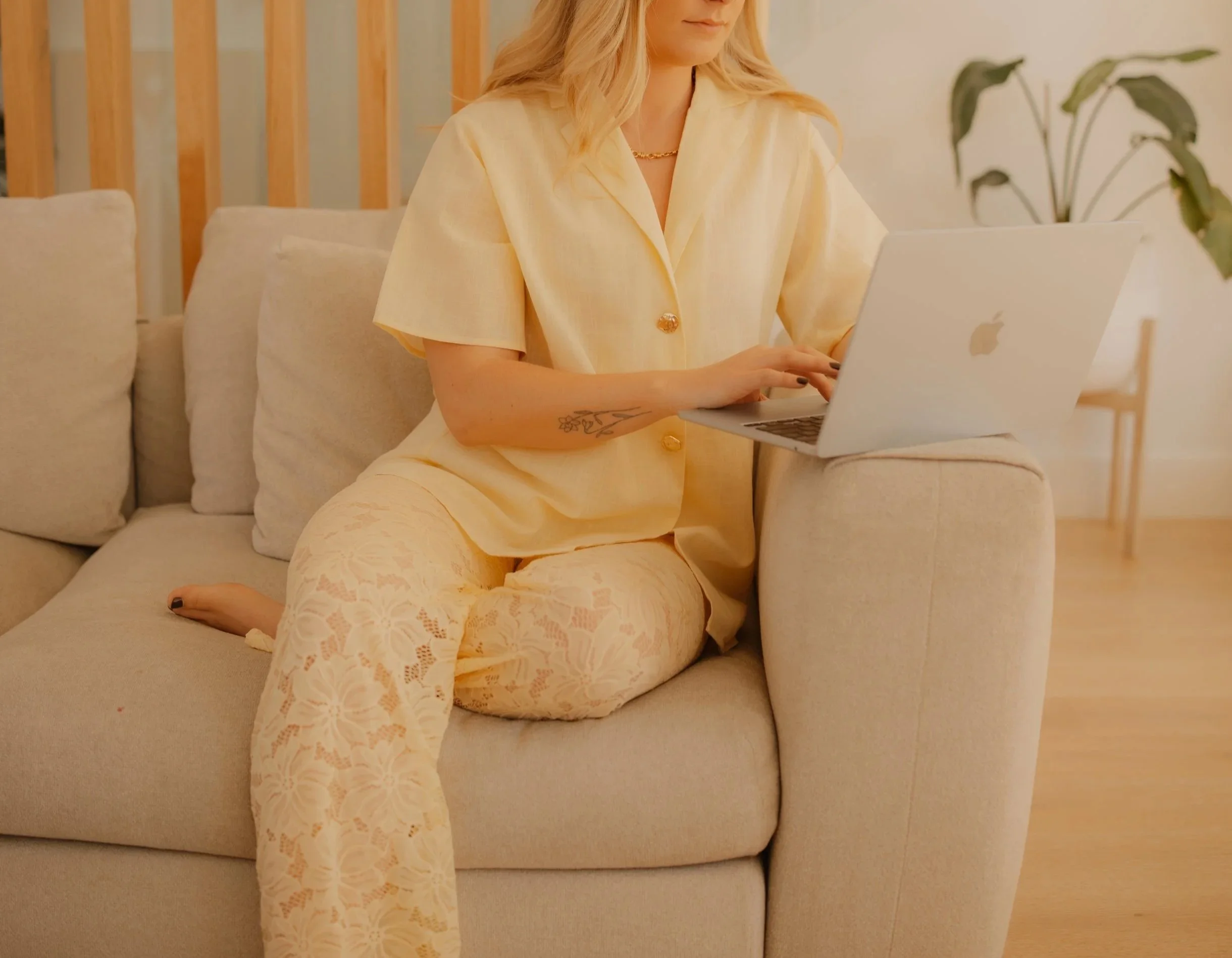 Woman wearing a yellow outfit sitting on a beige sofa, using a silver Apple MacBook, with a green plant in the background.